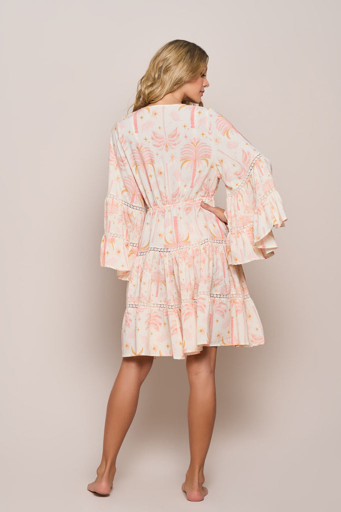 A model casually posing, showing the back view of the Barbie Short Kimono in white and light pink floral patterns, with embroidered patterns on the ruffles both on the bottom and on the hand flair, with a tie up belt, a perfect coverup for the same colored bikini top and bottom.