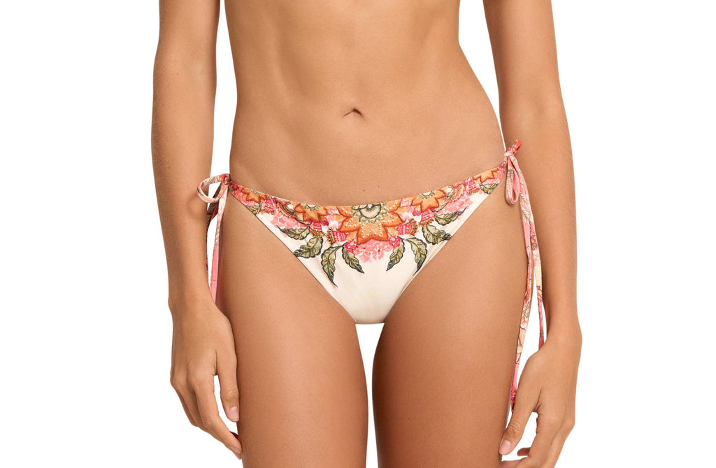 A close up picture of a model wearing Spirit Tie Side Bikini Bottom Cheeky, with adjustable tie side and a cheeky bottom coverage, colored beige or nude, with pink flower decorations.